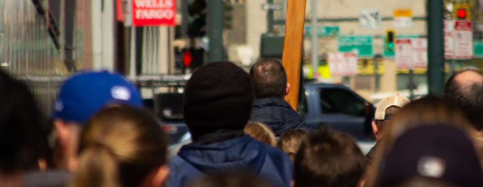A wooden cross is carried through a busy downtown street as a crowd follows behind in prayer.