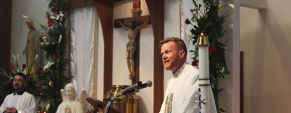 Priest in white robe speaks at a decorated church altar with floral arrangements, candles, and statues; reverent atmosphere.