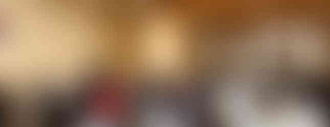 A crowded room with people standing and seated, facing a wall with engraved names and flower tributes. Wooden beams and a statue are visible.