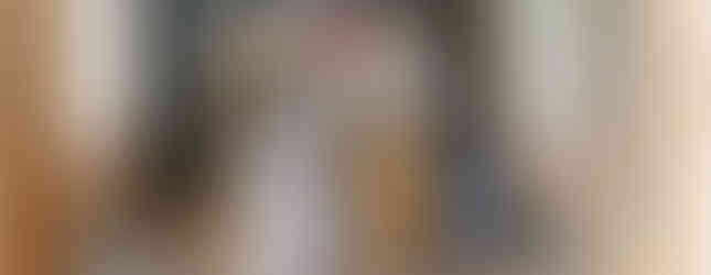 A person in clerical attire sprinkles holy water in a sunlit room with brick walls, a fan, and a clock. Quiet, reverent atmosphere.