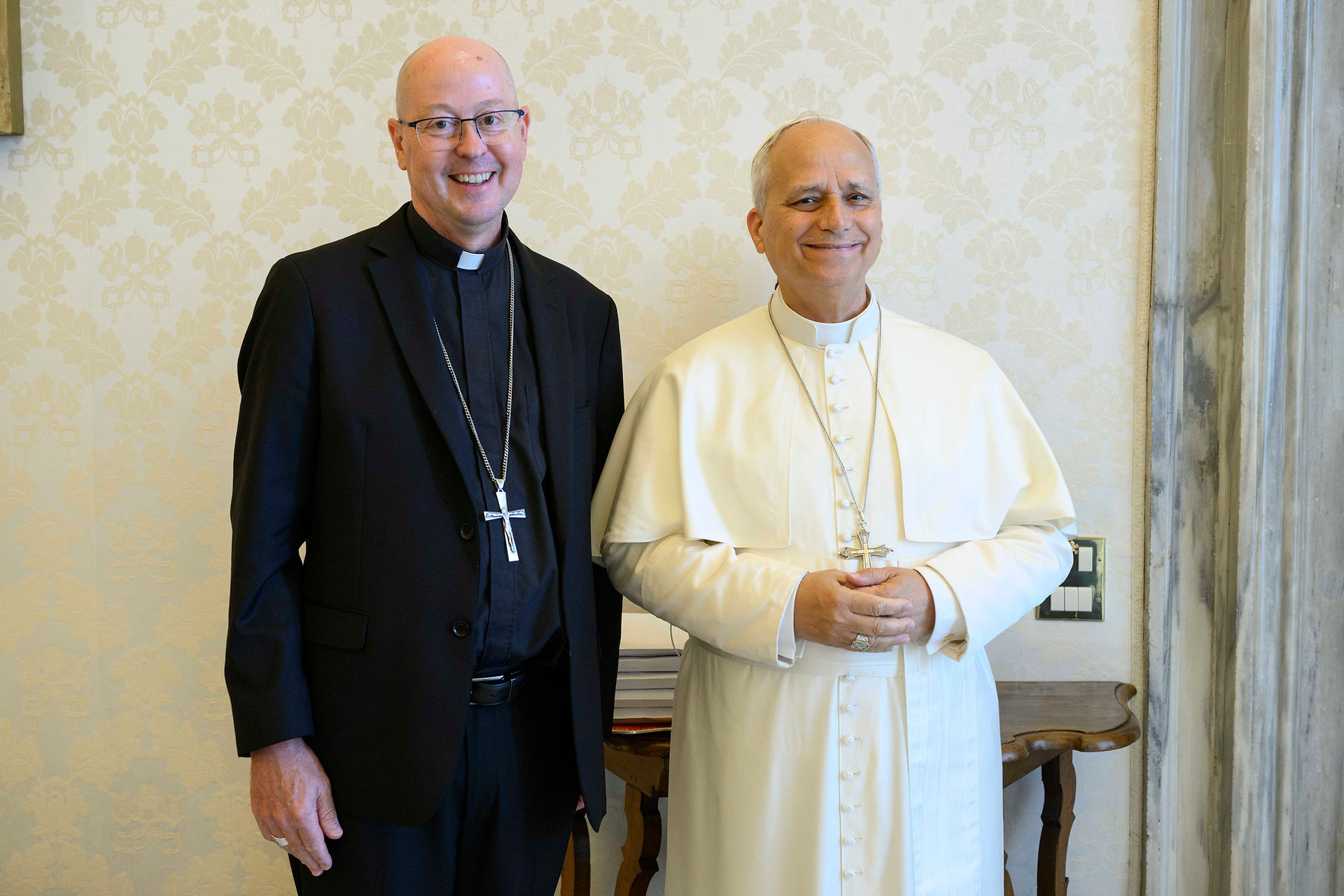 Two smiling clergymen pose indoors, one in black suit and one in white papal robes, before cream patterned wallpaper.