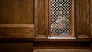 Man seen through a confessional screen in a wooden booth. The setting is dimly lit, conveying a solemn, contemplative mood.