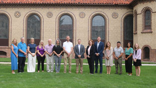 Twelve people stand in a row on a lawn before a brick building with arched windows, posing for a formal group photo.