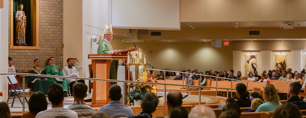 A bishop in green robes delivers a sermon at a wooden podium in a church filled with seated congregation. Religious icons adorn the walls.