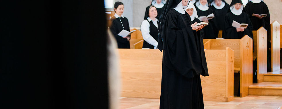 A newly professed sister reads from a prayer book during the liturgy while fellow sisters gather in choir behind her.