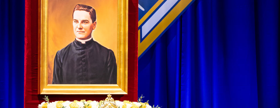 Portrait of a priest on a red pedestal with white roses, in front of a blue Knights of Columbus K of C backdrop