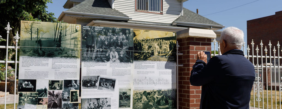 A man stands outside reading a large display board with historical photos and text mounted along a brick fence.