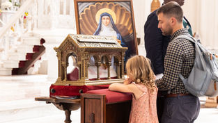 Family prays at a church reliquary beside an icon of a saint, with a man and little girl at the altar.