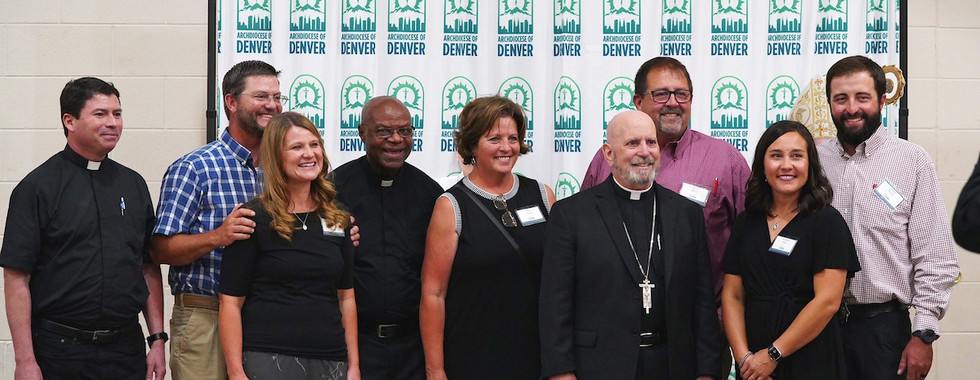 A group of smiling people, including clergy, pose in front of an "Archdiocese of Denver" backdrop in a hall. Bottles on the table.
