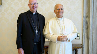 Dos clérigos sonrientes posan juntos: uno en traje negro y otro en blanco, con cruces, ante pared beige decorada.