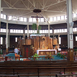 Spacious church interior with wooden altar and large crucifix, pews in foreground, and Jesus painted on white columns.
