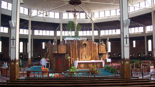 Spacious church interior with wooden altar and large crucifix, pews in foreground, and Jesus painted on white columns.
