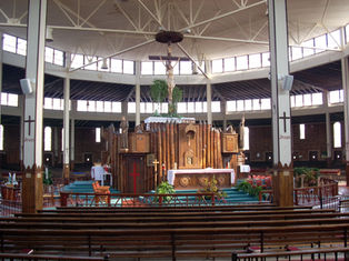 Spacious church interior with wooden altar and large crucifix, pews in foreground, and Jesus painted on white columns.