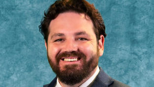 Smiling man with a beard in a blue suit and tie against a mottled teal background. The mood is positive and professional.