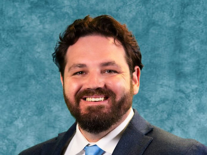 Smiling man with a beard in a blue suit and tie against a mottled teal background. The mood is positive and professional.