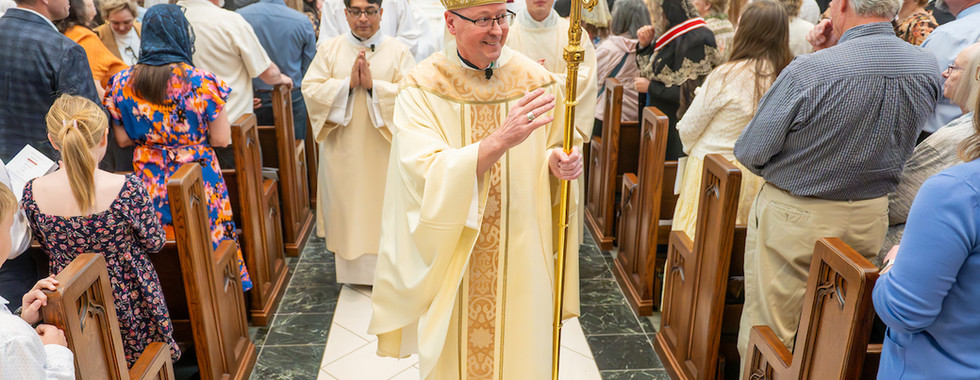 A bishop in ornate robes and mitre walks through a church aisle, holding a golden staff. A crowd in colorful attire looks on.