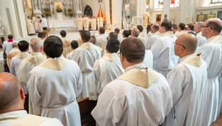 Priests in white robes with gold stoles gather in a cathedral during a ceremony. The altar, candles, and stained glass are visible.