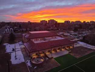A building with a red roof stands against a vibrant orange sunset, with snow patches and parked cars nearby in an urban setting.
