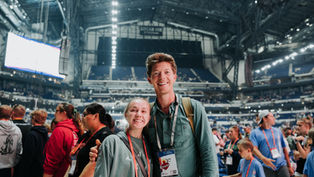 Smiling man and woman at a large indoor event with a crowd and bright lights in the stadium. They wear lanyards and casual clothes. Energetic mood.