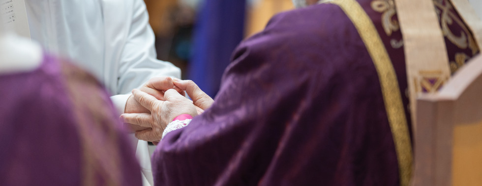 Two clergy members in purple and white garments clasp hands in a church setting, conveying a solemn and reverent mood.