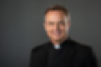 Clergy member wearing a black clerical suit smiles against a plain gray background, exuding a warm and welcoming demeanor.