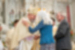 Bishop in ornate vestments shakes hands with a white-haired woman in blue inside a cathedral, with a suited man nearby.