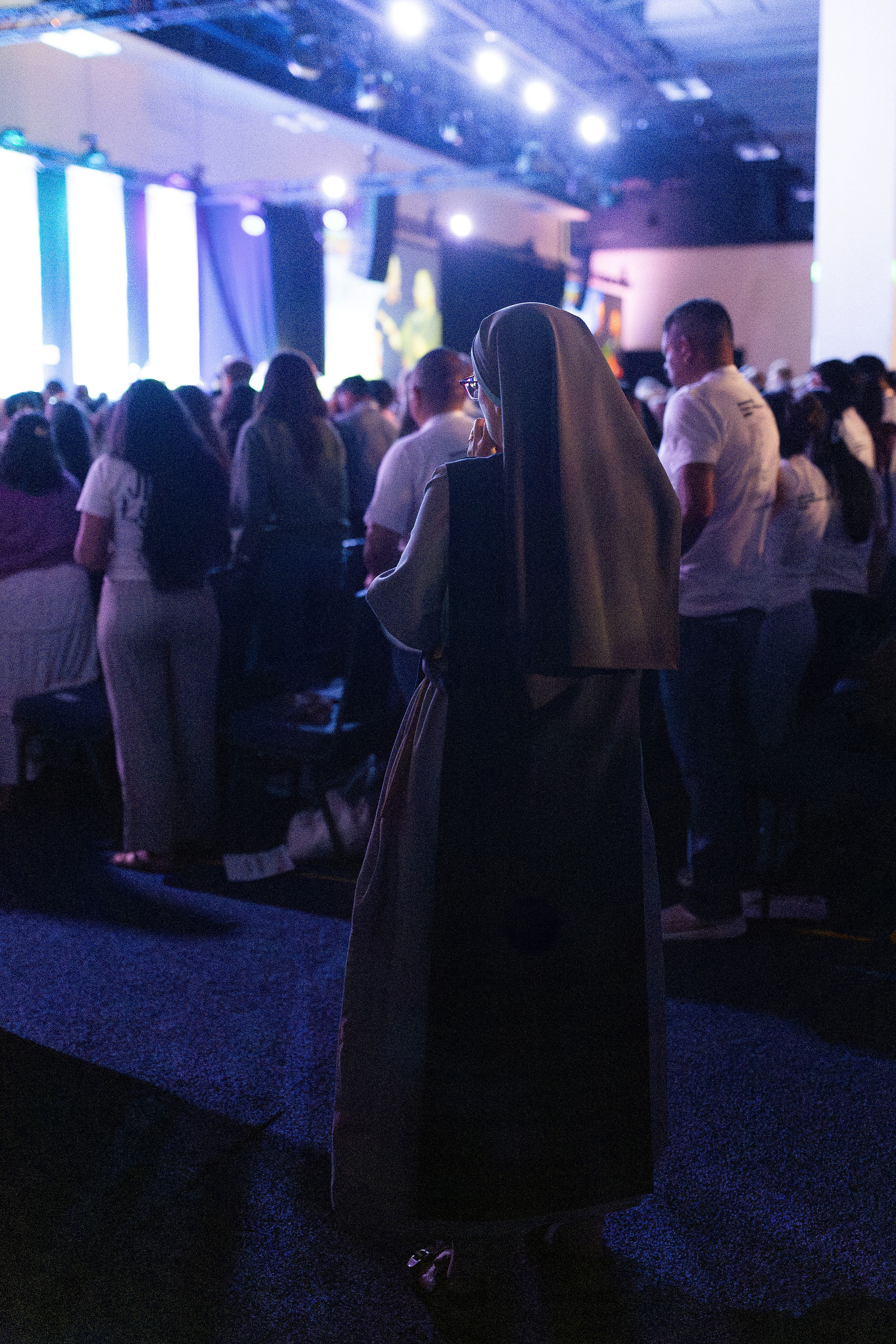 Nun in habit stands among a crowd at an indoor conference, facing a brightly lit stage with large screens and blue-purple lights.