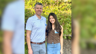 Man and woman smiling outdoors, man in patterned shirt, woman in brown vest with blue skirt. Sunlit leafy background. Both wear name tags.