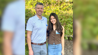Man and woman smiling outdoors, man in patterned shirt, woman in brown vest with blue skirt. Sunlit leafy background. Both wear name tags.