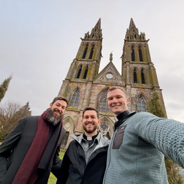 Tres hombres sonrientes posan frente a una iglesia de piedra con dos torres altas. El cielo está nublado, dando un ambiente sereno.