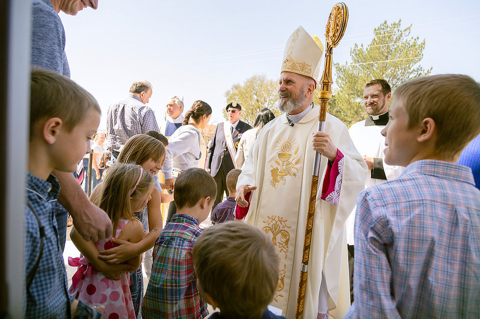 Bishop in ornate robe with staff greets children and adults outdoors on a sunny day. People wear colorful outfits, creating a joyful scene.