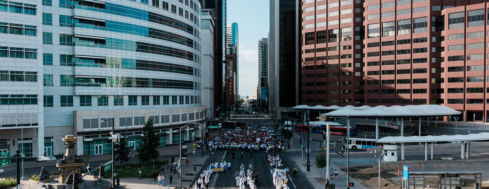 Multitud en desfile por una avenida entre rascacielos; se ven The Denver Post y BOK Financial bajo cielo despejado.