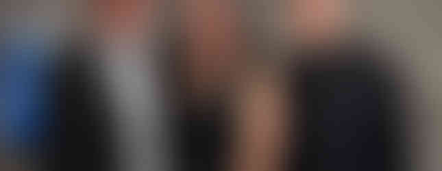 Three people smiling at a formal event, wearing suits and a sleeveless dress. A blue-lit floral arrangement in the background.