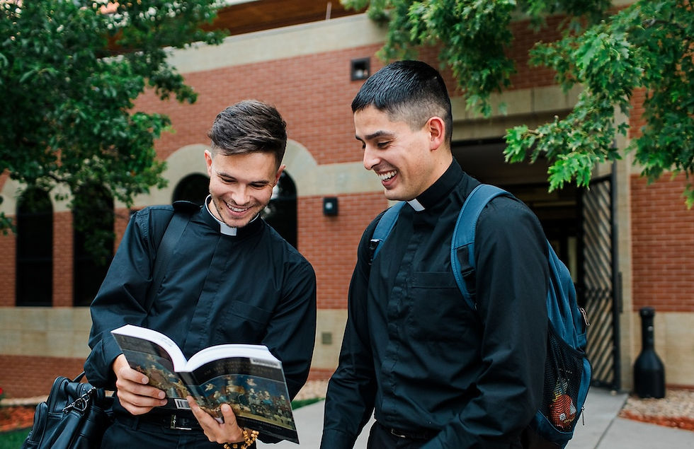 Two men in black clerical clothing smile while reading a book outdoors against a brick building and green trees. One carries a backpack.