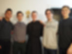Five young men smiling and standing shoulder to shoulder indoors. One wears a University of Denver hoodie. A cross hangs on the wall behind them.