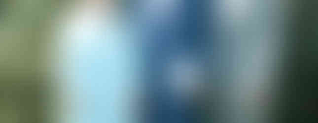 Three people pose in front of green bushes. A graduate in a blue cap and gown holds a diploma, flanked by two adults in formal attire.