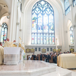 Catholic mass in a bright cathedral, priests at the altar and congregation seated beneath tall stained-glass windows.