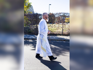 A person in a white robe walks on a sunny street, holding a rosary. Background includes a fence, trees, and distant buildings.