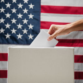 Hand placing a ballot into a box against the backdrop of a U.S. flag, symbolizing voting and patriotism.