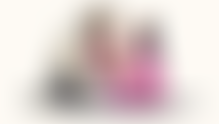 A bearded man in white, a woman in pink, and a baby in a purple wrap share an emotional moment on the ground, conveying tenderness.