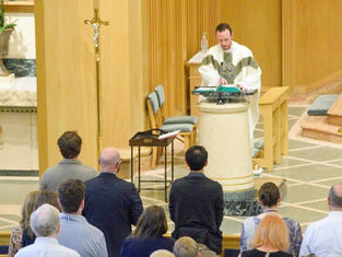 Priest in white robe speaks at a podium in a church. Congregation stands, facing altar. Cross and plants visible. Calm atmosphere.
