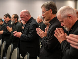 Men in black suits and clerical collars stand in prayer with heads bowed and hands clasped, in a softly lit room, creating a solemn mood.