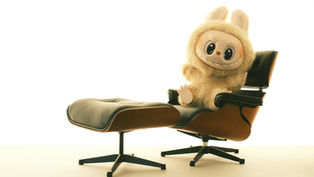 Cute plush toy with rabbit ears, smiling on a black leather chair with a matching footrest. Minimalist white background.