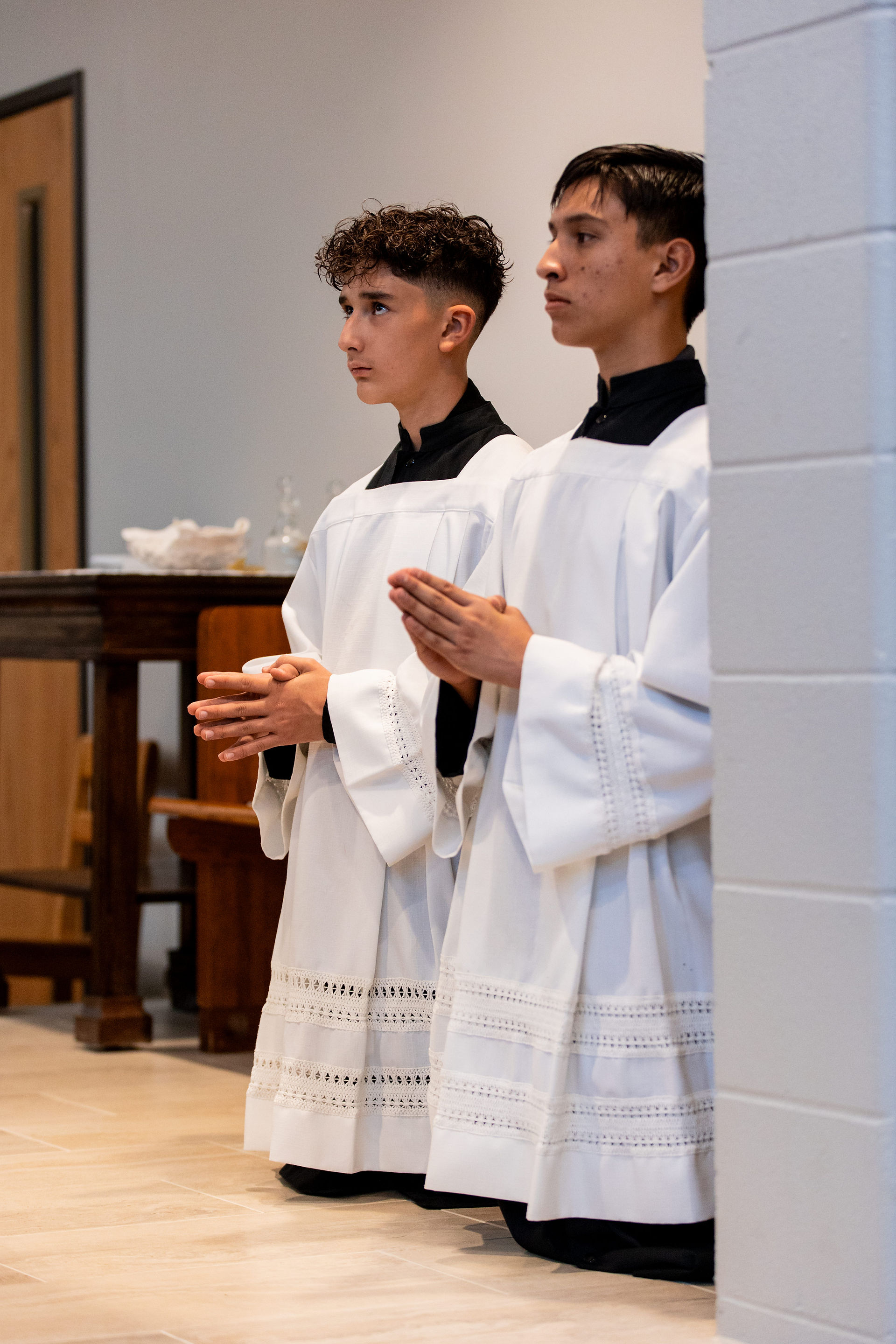 Two altar boys kneel in prayer, wearing white robes with lace detail. They're in a church setting with wooden furniture visible.