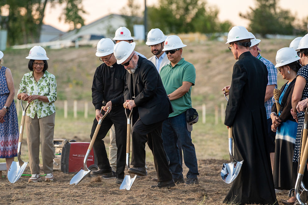 People in hard hats participate in a groundbreaking ceremony, using shovels on a grassy field. Sunny background, some are smiling.