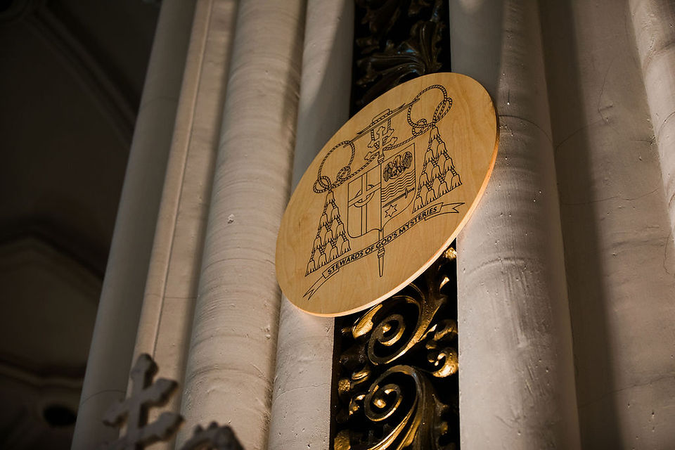 Escudo episcopal tallado en madera colocado entre columnas dentro de la iglesia.