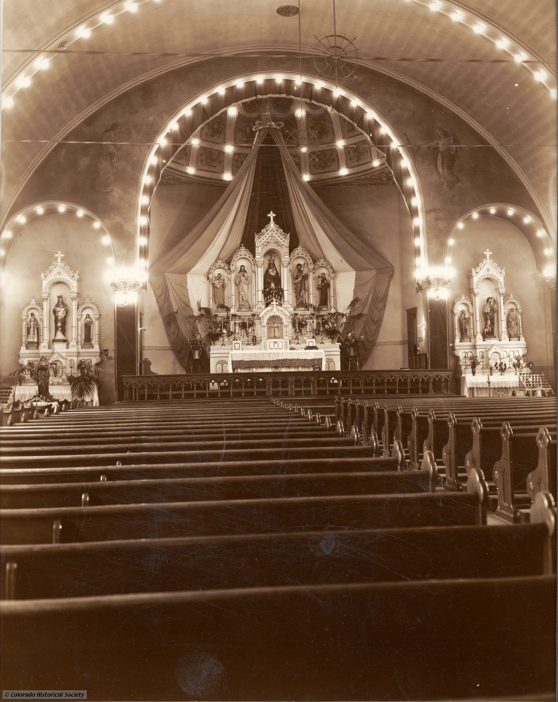 Sepia photo of an empty church nave with wooden pews facing an ornate altar framed by lights; Colorado Historical Society watermark.