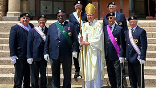 Bishop in gold vestments poses with seven uniformed men on church steps, holding a crozier; all smiling.