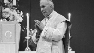 Bishop in white vestments stands in church, hands clasped in prayer beside lit candles and flowers, solemn black-and-white scene