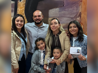 Family of six smiling in front of a colorful mural. One holds a trophy, another a photo ID. Casual attire, warm atmosphere.